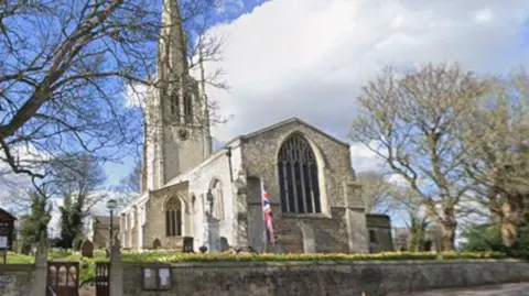 A parish church in what appears to be a rural village. The church grounds are marked by a long stone wall. A union flag stands in front of the church on a flagpole. It is a sunny day.