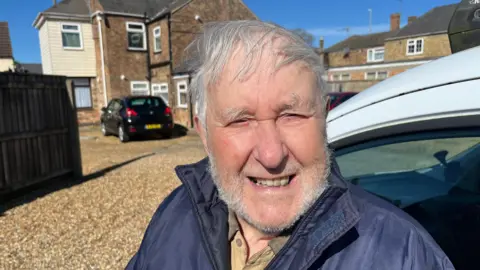 John Devine/BBC Colin Bedford, 89, has grey hair swept across his head, and a grey stubbly beard and moustache. He is wearing a blue coat with a brown shirt underneath. Behind him is a house with some cars parked on a gravel driveway.