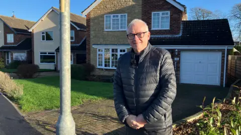 John stands to the right of the lamp-post in front of housing. A driveway is right next to the lamppost.
