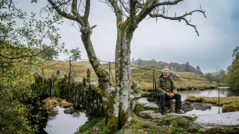 Colin Hindle Colin is sitting on some stones beside a stone bridge and river. He is staring out into the distance. There are green hills in the background and a tree is beside him.