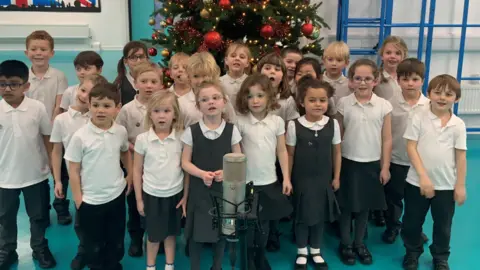 A group of young children wearing black, white and grey school uniform standing behind a microphone with a Christmas tree behind them.