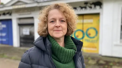 Carrie Sage stands outside of Bristol Zoo Gardens. She has short mousey brown hair and wears a dark-green turtleneck underneath a navy-blue puffer jacket.