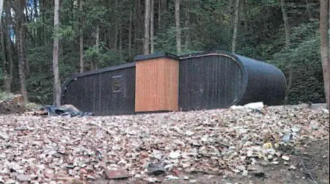 Just in front of many trees in a woodland, a black oval-shaped accommodation pod sits in a clearing. The floor is covered in bricks and rubble.