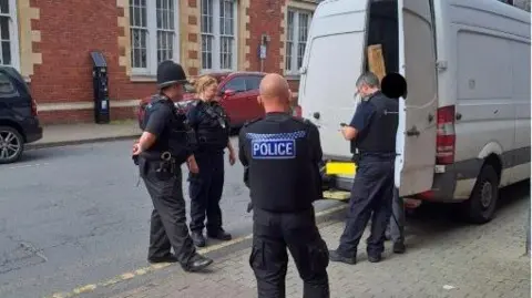 Four police officers, three men and one woman are standing on the street next to a white van. There is a black circle over someone's head, who has been arrested. One of the back doors of the van are open.