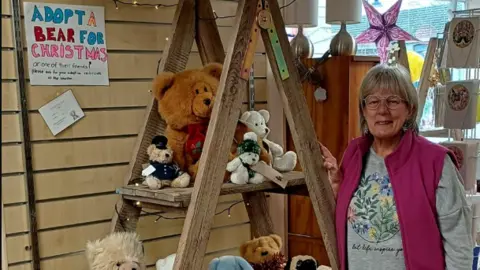 St Margaret's Hospice Woman in pink gilet with teddy bears and a sign which says 'adopt a bear for christmas'