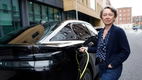 Ford UK's managing director Lisa Brankin wearing blue jeans, a navy blue blouse with white polka dots and a navy blue cardigan. She is standing next to a black Ford Capri connecting it to an electric charger and smiling at the camera