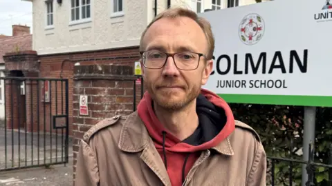 Cameron Noble/BBC Man with glasses, a hoodie and a brown coat stands outside a mostly brick-built school. There's a sign saying Colman Junior behind him. 