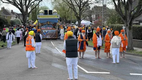 Image shows a residential road filled with hundreds of people, in the middle is a decorated lorry which the crowd is close to on either side. In the foreground of the image are a group of Sikh men dressed in traditional orange clothes and turbans. 