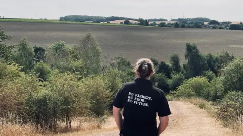 Paul Rowland A woman is stood overlooking acres of fields and countryside with her back to the camera. She has dark hair pulled back and is wearing a black t-shirt which says "no farmers, no food, no future"