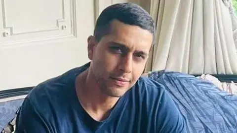 Picture of Kamran Aman wearing a blue t shirt. He is looking straight at the camera. He has short dark hair and is clean shaven. 