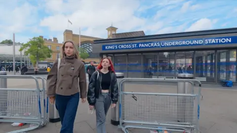 Two women walk past the entrance to King’s Cross St Pancras Underground Station in London. Metal barriers line the pavement in the foreground, while the station building and a clock tower are visible behind the blue station sign under a partly cloudy sky.