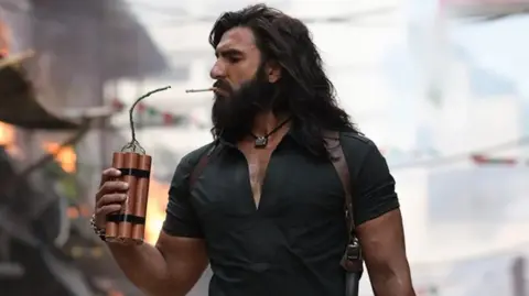 Ranveer Singh in a still from Dhurandhar, seen holding dynamite to light a cigarette. He's wearing a black shirt with a silver pendant around his neck. He has long hair and a beard.
