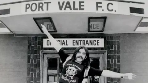 IFK Legacy CIC A black and white photograph of Motörhead frontman Lemmy, a man with long dark hair and a dark moustache, holding his arms out like a plane underneath a sign reading "PORT VALE F.C.". Another sign under that reads "OFFICIAL ENTRANCE".
