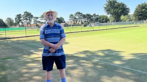 BBC Organiser Chris Holt stands in front of a link fence inside of which are lots of tennis courts marked out with white lines and mesh nets. Chris wears a blue horizontally striped t-shirt, dark blue shorts and blue socks. It's a very sunny day so he has a sun hat on to shade his face. He has white hair and beard. 