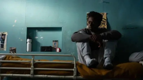 James A Demetriou A man sits on the top bunk of a prison bed, backed by a patchy blue wall. He has his eyes closed.