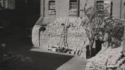 Essex Police A ladder resting against several sandbags placed in front of a police station.
The sandbags have been stacked to the first window. The image is in black and white.