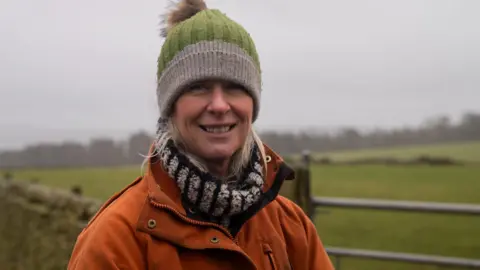 Kathy Weir is wearing a thick orange coat and a green and grey bobble wool hat. She is smiling and there are green fields behind her