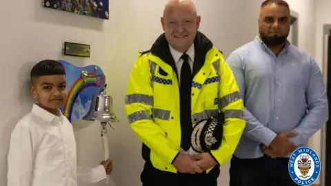 Abu Bakr with Chief Constable Craig Guildford and his dad PC Mohammed Azam as he rings the bell to mark the end of his inpatient cancer treatment. Abu Bakr wears a white shirt and holds the bell's string in his left hand and looks at the camera. The bell has a rainbow and clouds picture next to it. Mr Guildford in the centre is wearing a black tie and black trousers with a green police jacket and is holding his hat with his hands folded. PC Azam wears a blue shirt and has his hands folded in front of him. 