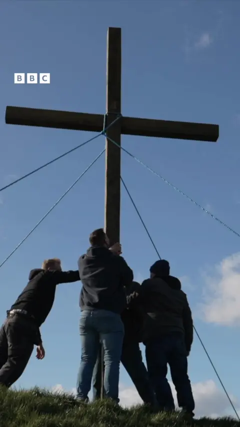 A wooden cross is put up on a hill. Three people's silhouettes are in the foreground.