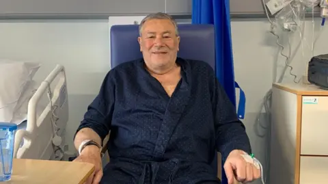 Gordon Anderson/BBC A man sitting by a hospital bed in a ward. He is wearing a blue dressing gown and has a cannula in his left hand. He is smiling as he looks at the camera. 