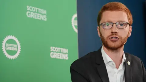 PA Media Ross Greer, who has ginger hair and glasses, pictured in front of Scottish Green branding. He is wearing a dark suit jacket and white shirt. 