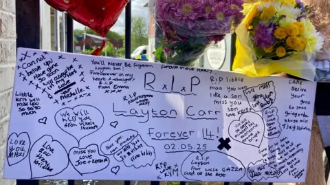 A large banner inscribed R.I.P. Layton Carr, Forever 14, with a number of signed notes and drawn hearts, paying tribute to the teenager. It is pinned to a gate and flowers and a balloon can be seen above it.