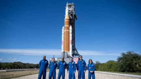 A huge orange-and-white Moon rocket stands upright on its launch platform against a cloudless blue sky. In the foreground, seven astronauts in bright blue flight suits line up on a pale gravel roadway, facing the camera. An American flag hangs from the launch structure behind them, with low trees and flat grassland stretching away on either side.