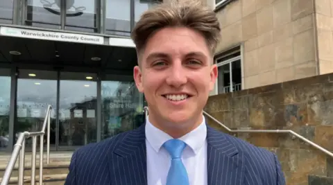 A young man with short light brown hair is wearing a blue pinstriped suit, a white shirt and pale blue tie. In the background are steps leading up to a large glass building with a sign above double doors that reads, "Warwickshire County Council".