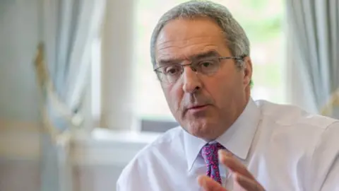 McPhillips A man with grey hair and glasses in a white shirt with a pink patterned tie, in front of a blurred image of a room