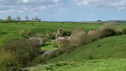 A wide rural landscape of rolling green hills under a bright sky filled with scattered white clouds. In the middle distance, partially hidden by trees, sits the stone ruins of an old church. The surrounding fields stretch far into the horizon, dotted with trees and hedgerows, creating a peaceful countryside scene.