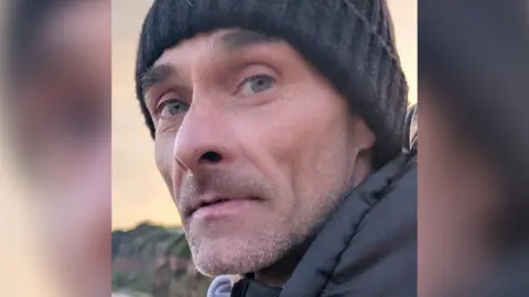 Missing man Jason. He is wearing a dark looking woolly hat and is looking at the camera. He has grey stubble and is wearing a dark coat.