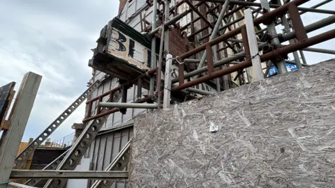 Martin Heath/BBC Close-up of a heavily scaffolded building with timber hoarding around the base, showing damaged brickwork and part of an old shop sign protruding through the metal framework.
