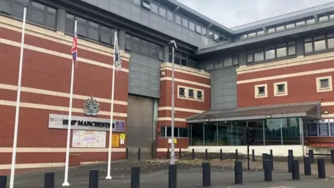 A view of the front of Strangeway prison, including the prison sign, flag poles, and the glass panelled entrance way by the metal door for vehicle entry.