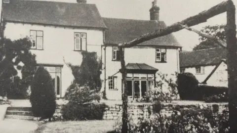 Guide Dogs A black and white image of an old white wall building from its back garden with black framed windows and dark sloping roofs. A patio area steps down into a garden, with flowers, shrubs and hedges.