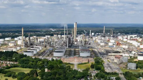 ExxonMobil Oil refinery site with multiple tall chimneys and numerous large round containers with green fields on its outskirts.