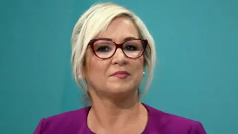 Michelle O'Neill - she has blonde hair and is wearing a purple top and red-rimmed glasses.