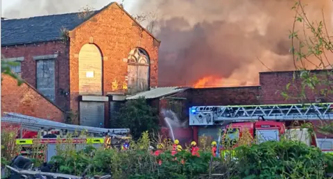 Fire tears through a red-brick building as smoke plumes. A fire engine is in the foreground and crews can be seen spraying water.