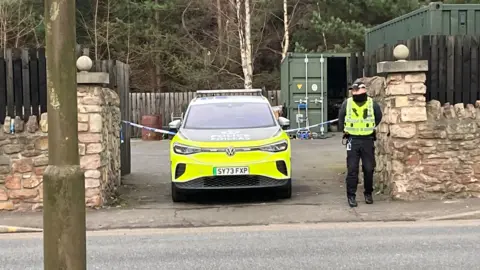 A uniformed police officer stands next to a police car which is parked in a driveway which has been taped off