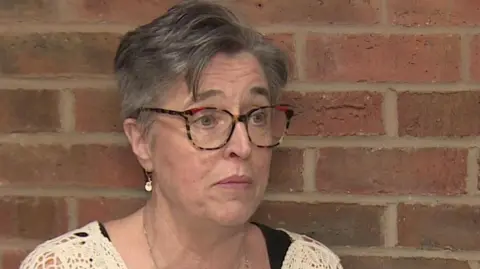 Gill Gray. She has short grey hair and is wearing large glasses. She is wearing a loosely knit beige cardigan.