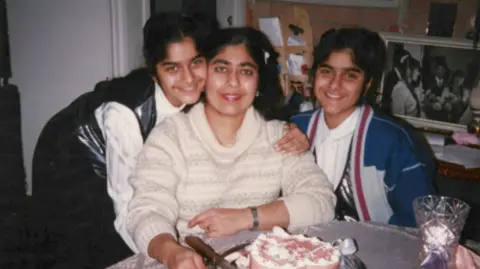 Chaudhry family A family photograph of Annsa Chaudhry, wearing a white jumper and sitting at a table with a birthday cake on it, with her daughters either side of her.