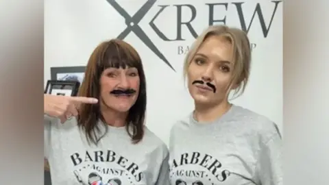 Somerset Live Two female barbers with fake moustaches wearing company uniform of a grey t-shirt with a poster for their shop, Krew Barbers, in the background