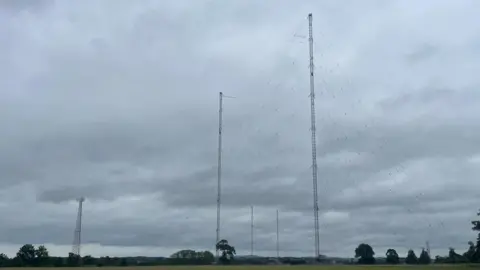 Five tall transmitter masts with wires between them and coming down to the ground are visible against a grey sky. On the ground is a green field and several green trees.