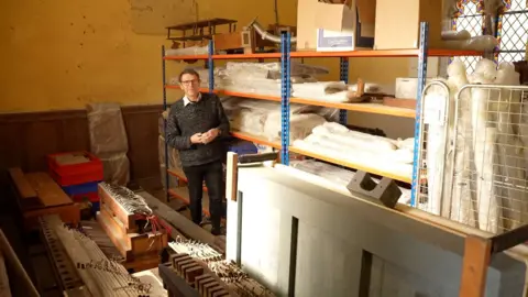 George Allan of Pipe Up standing by the remains of organs he has saved. There are shelves full of material and several wooden objects on the floor.