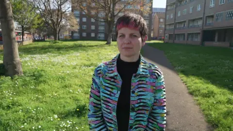 Zoë Garbett in a park with trees and grass behind her along with surrounding blocks of flats