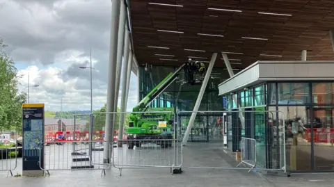 Temporary metal fencing across the entrance to a building, with a man in hi-viz stood to the side. There is a raised cherry picker under the building's canopy