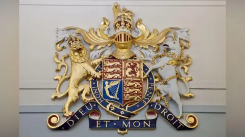 The Royal Coat of Arms of the United Kingdom, symbolising that justice is carried out in the name of the Crown. It is displayed prominently in magistrates' courts.