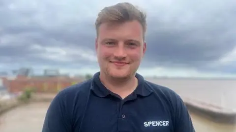 Alistair Sceats smiling into the camera and he is wearing a navy t-shirt with the Spencer logo on the front. He has short, fair hair and is standing in front of the Humber Estuary.