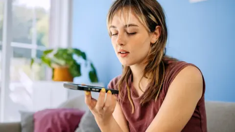 A young woman casually seated on a sofa, holding her smartphone and sending a voice message.