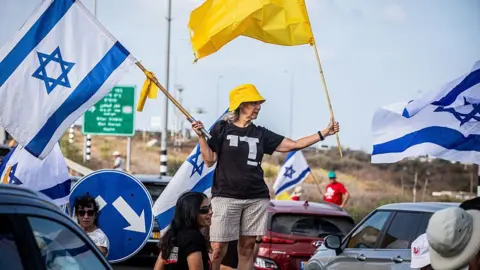 A woman holding an Israeli flag and a yellow flag representing the hostages waves them while stood in the middle of a busy road at a daytime protest. 