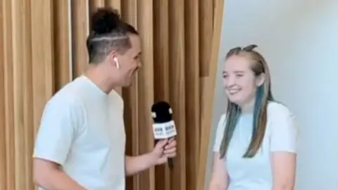 A BBC reporter with a bun talking to a girl with blue and blonde hair
